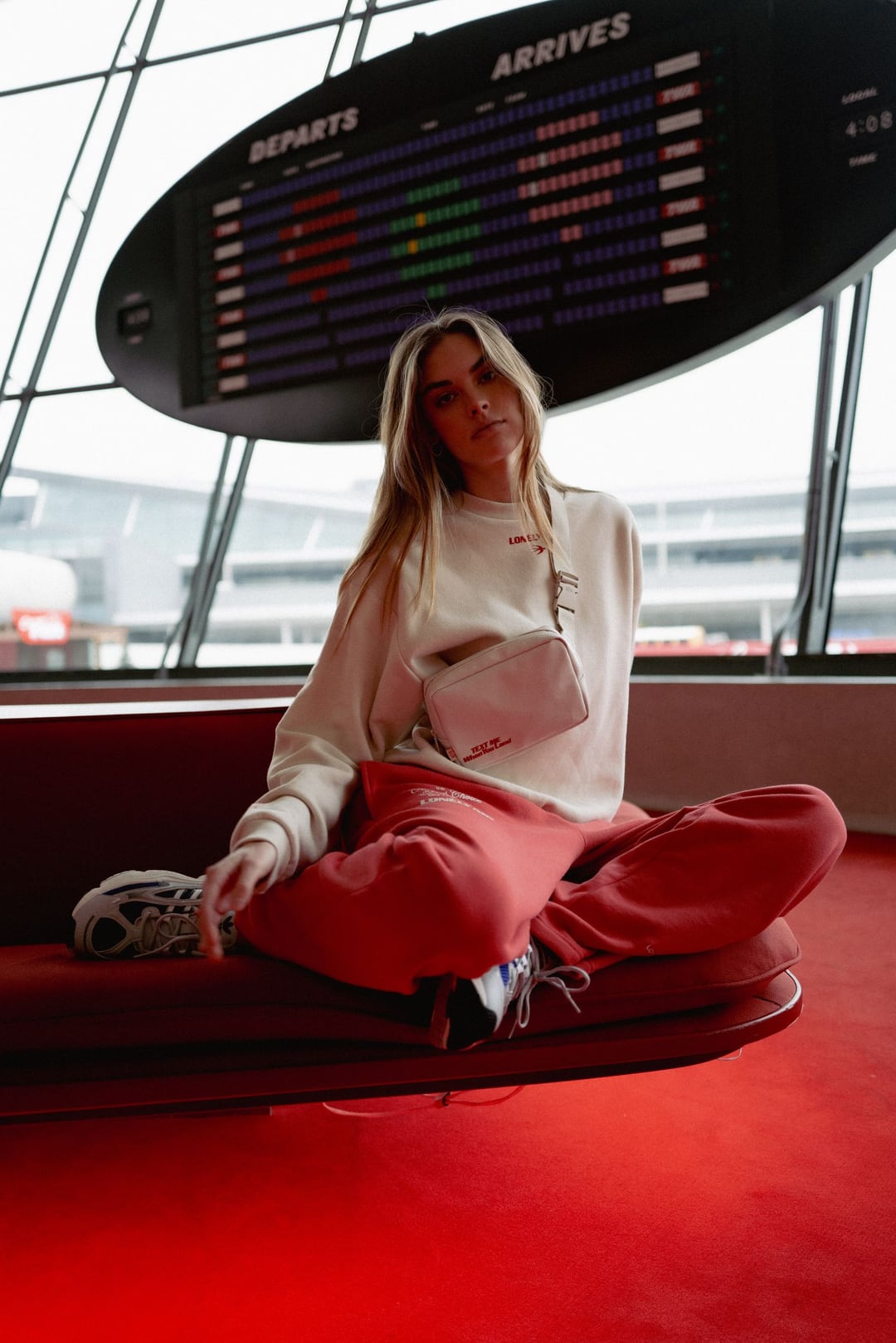 A model sits cross-legged on a red sunken lounge beneath a split-flap departures board, cream sweatshirt and red trousers.