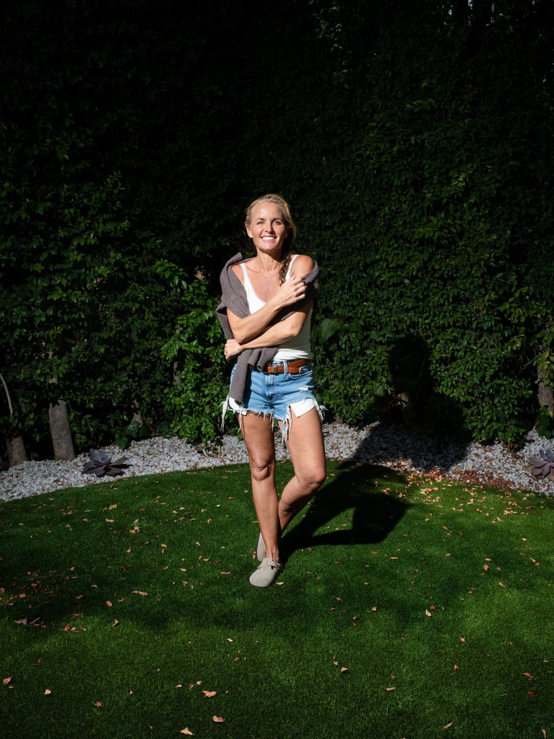 Davinia Taylor standing on a lawn against a dark hedge in hard sun, arms crossed, a grey sweater over her shoulders and denim cutoffs.