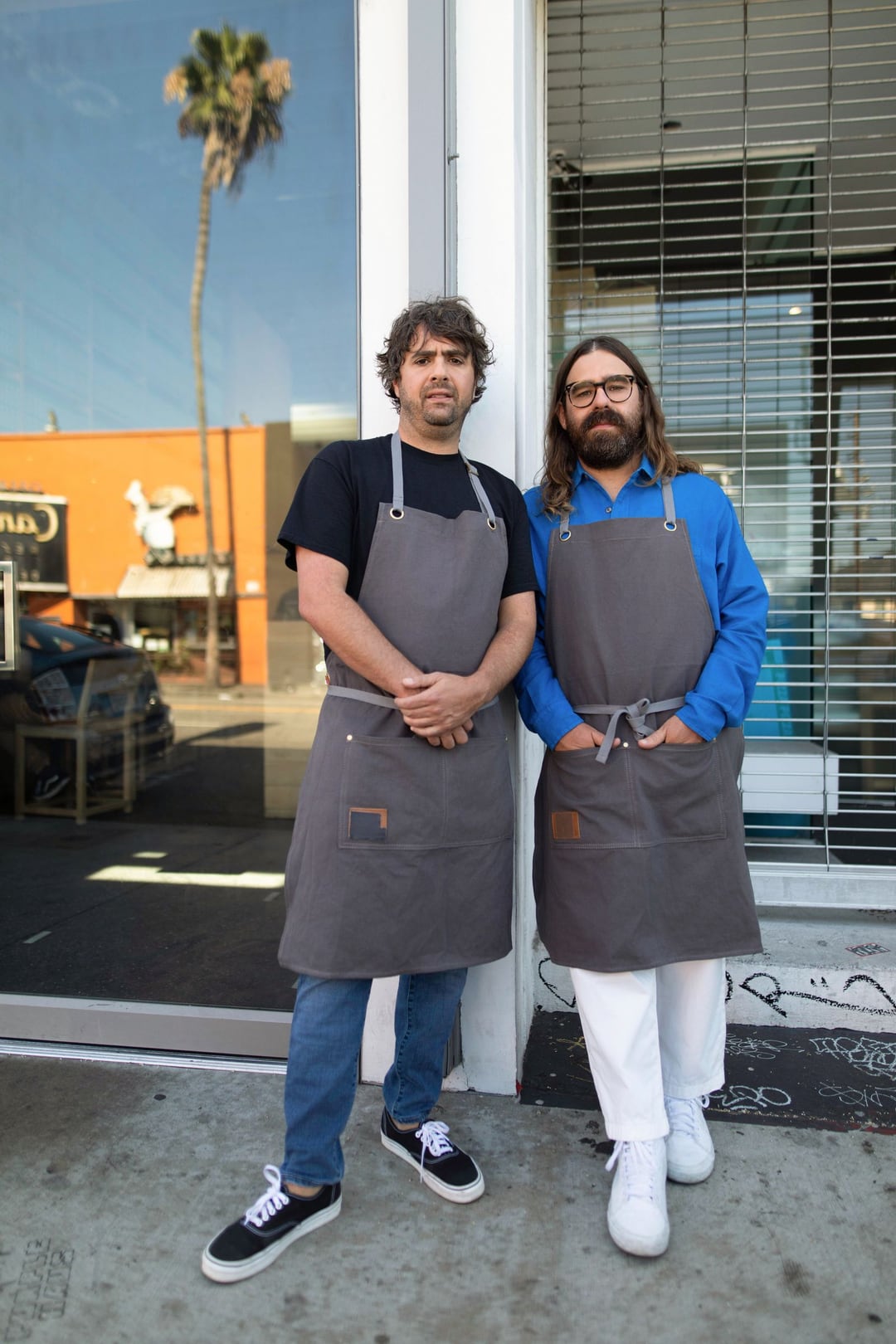 Jon Shook and Vinny Dotolo standing in grey aprons outside a shopfront window, a palm tree and an orange building reflected in the glass.