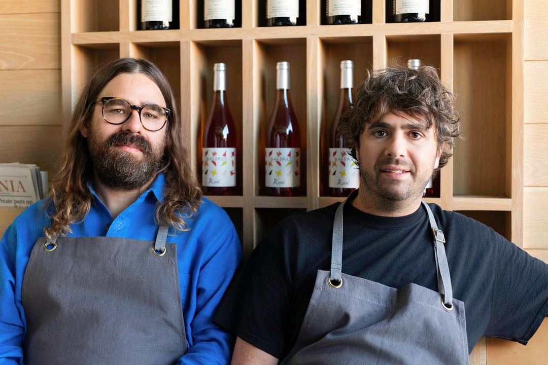 Chefs Jon Shook and Vinny Dotolo side by side in grey aprons, one in glasses and a cobalt shirt, in front of a wooden rack of wine bottles.