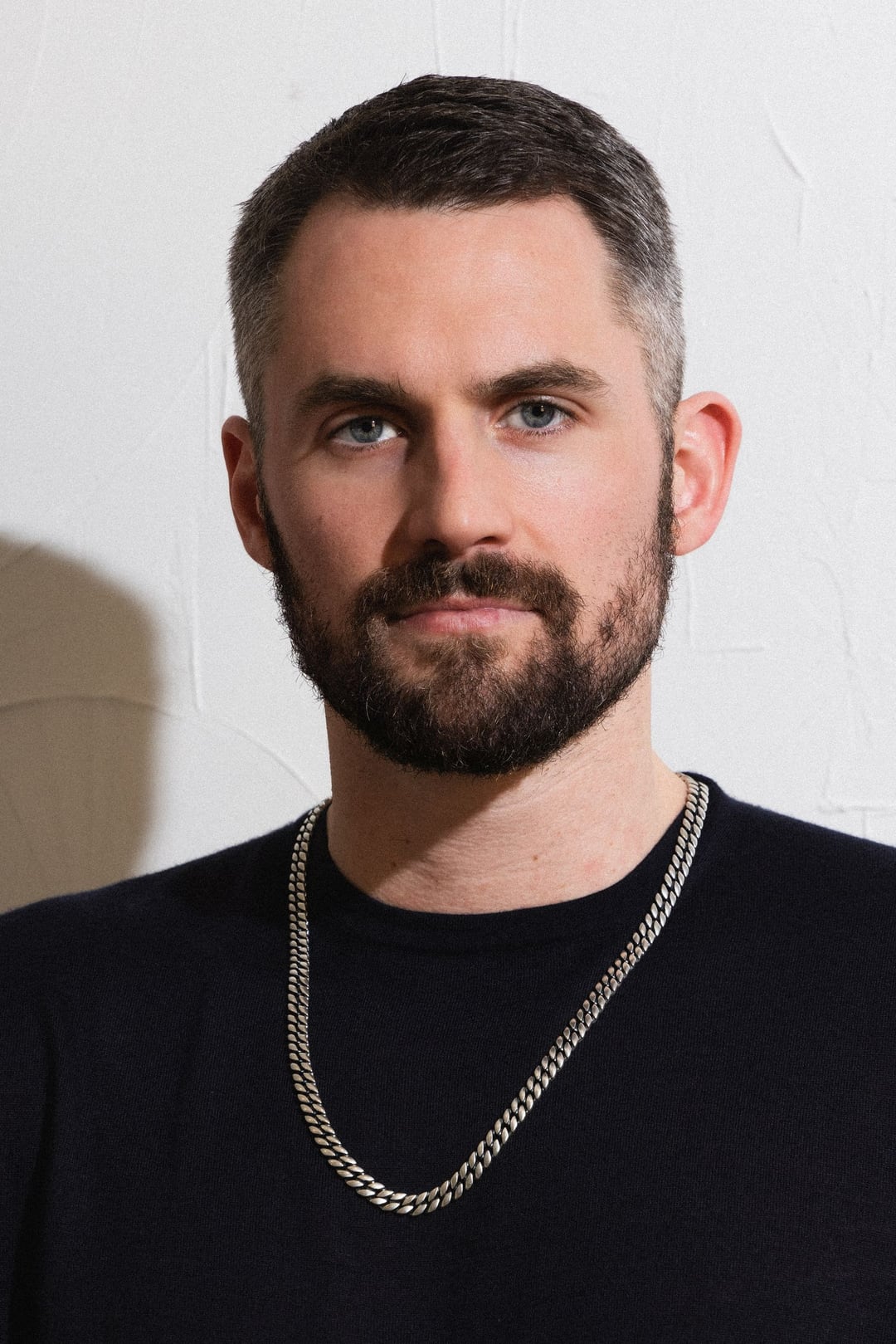 Kevin Love close-up against a white wall, black tee and a heavy silver chain, looking straight into the lens.