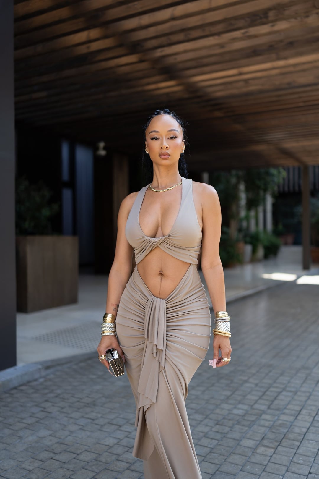 Draya Michele walking toward camera in a draped taupe two-piece under a slatted wood canopy, gold cuffs on both wrists.
