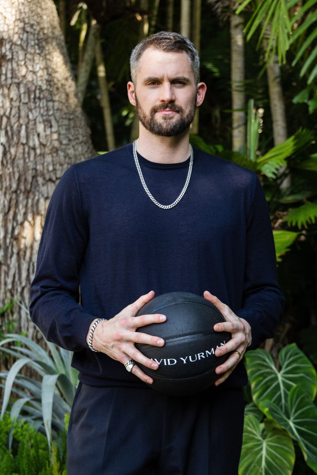 Kevin Love in a navy knit holding a black David Yurman basketball among tropical leaves and palm trunks.