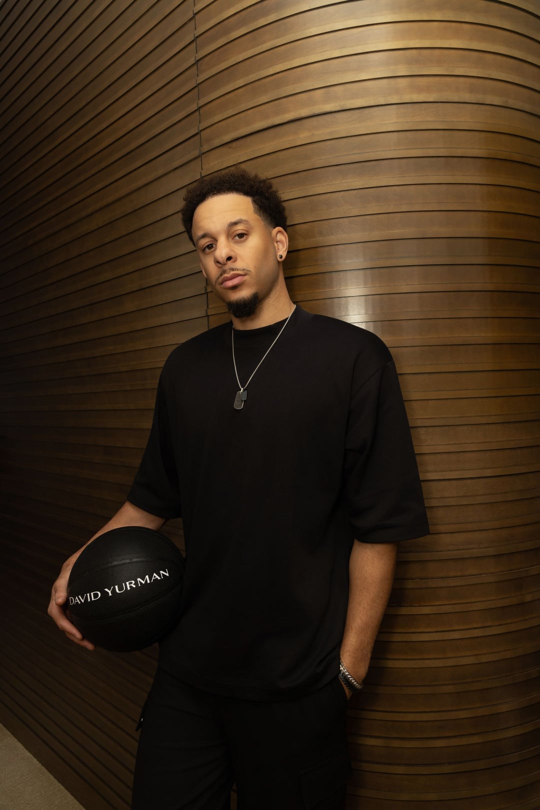 Seth Curry in black leaning on a curved bronze-slatted wall, holding a black basketball at his hip.