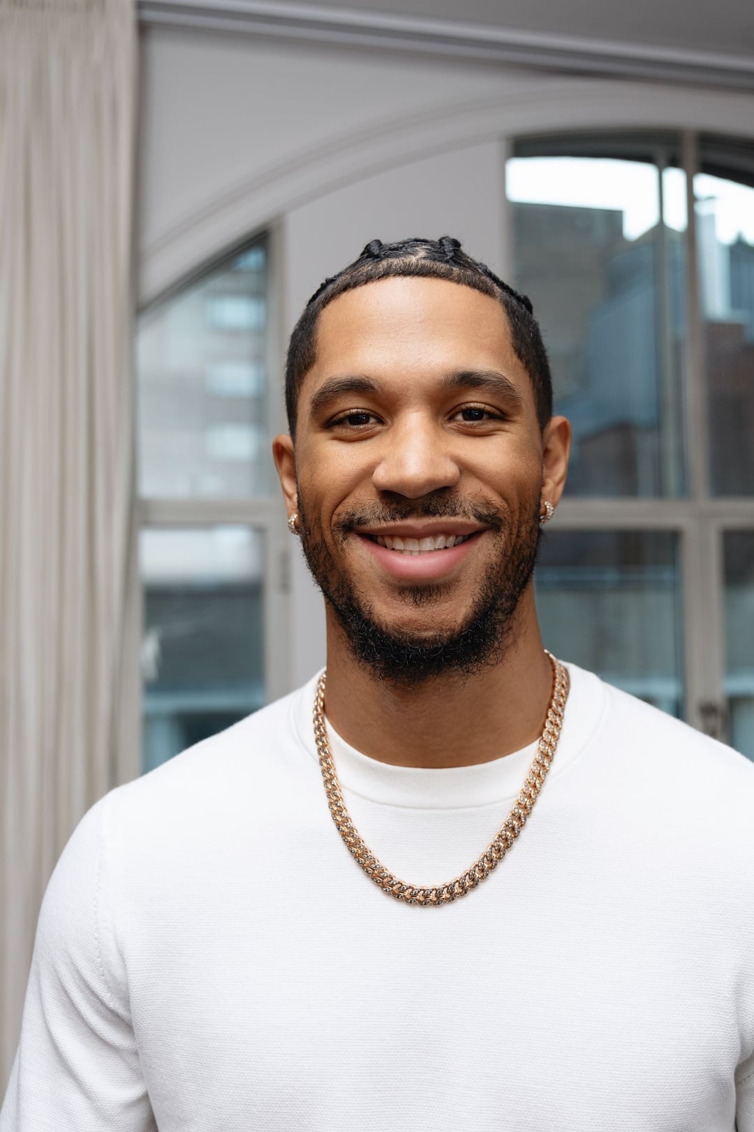 Josh Hart smiling close-up in a white knit and gold chain, arched windows behind him.