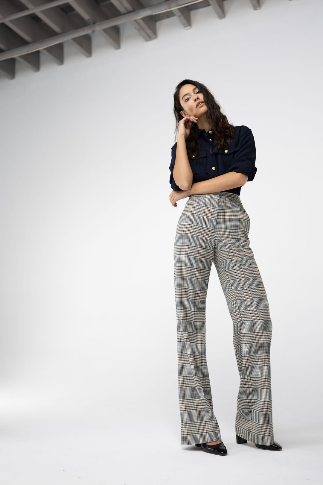 Model in a navy blouse and wide plaid trousers standing in a white-walled studio, hand to her hair.