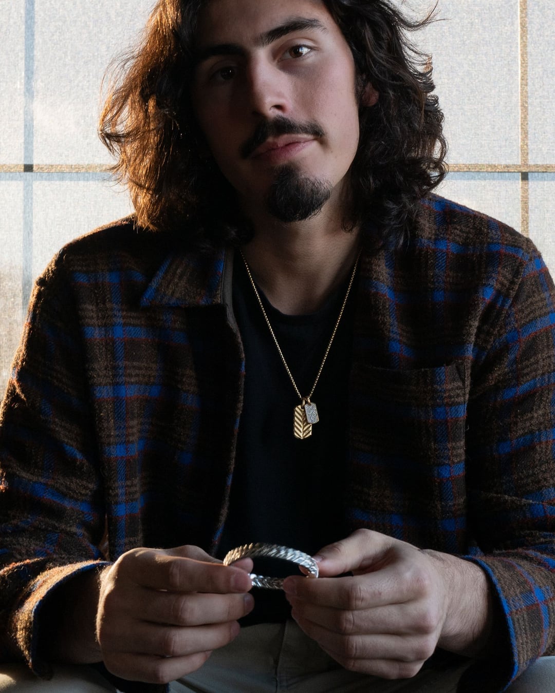 Jaime Jaquez Jr. seated in a plaid overshirt, turning a silver bracelet in his hands, looking straight at camera.
