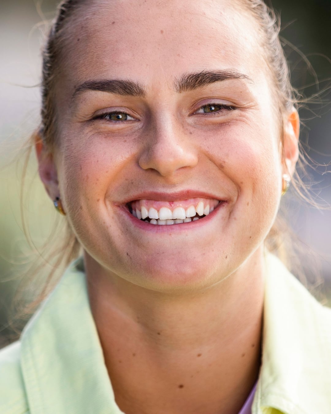 Aryna Sabalenka close-up, laughing into the lens in a pale yellow jacket, her hair pulled back.
