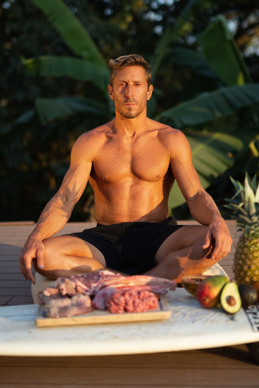 Paul Saladino shirtless and cross-legged on a deck in low golden light, raw meat on a board, a mango, avocados and a pineapple on a surfboard in front of him, banana leaves behind.