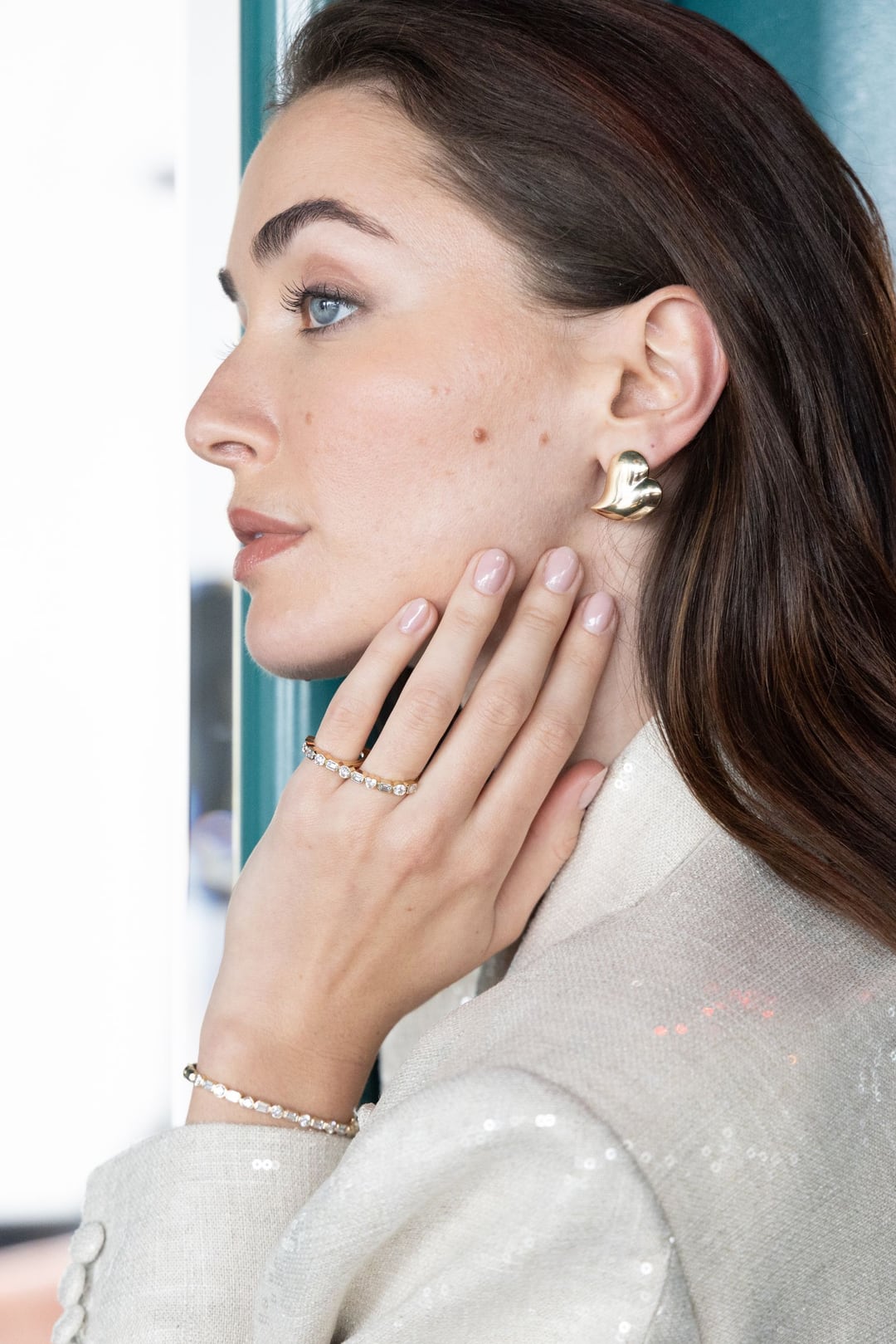 A model in profile against a window, one hand at her neck, gold rings and a sculpted gold earring, a cream sequinned jacket.