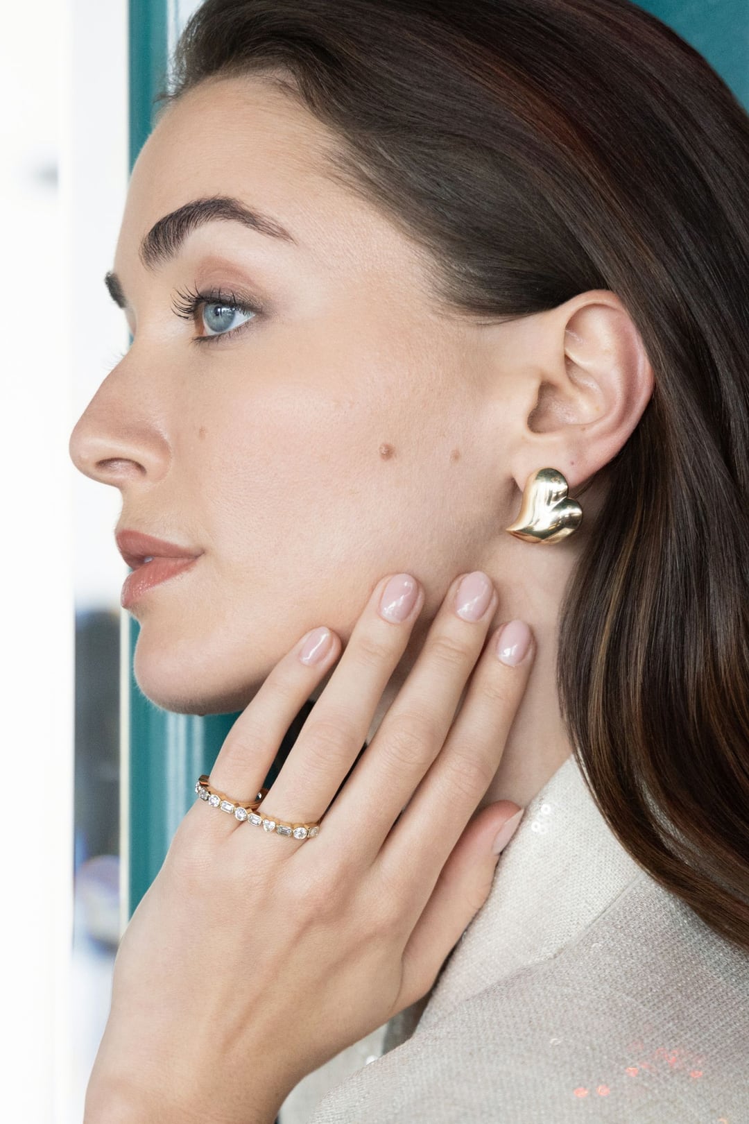 A model in profile against a window, one hand at her neck, gold rings and a sculpted gold earring, a cream sequinned jacket.