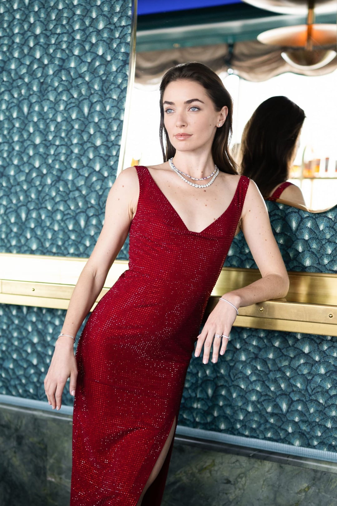 A model in a long red dress and a diamond necklace leaning against a teal bar, three-quarter length, a mirror behind her.