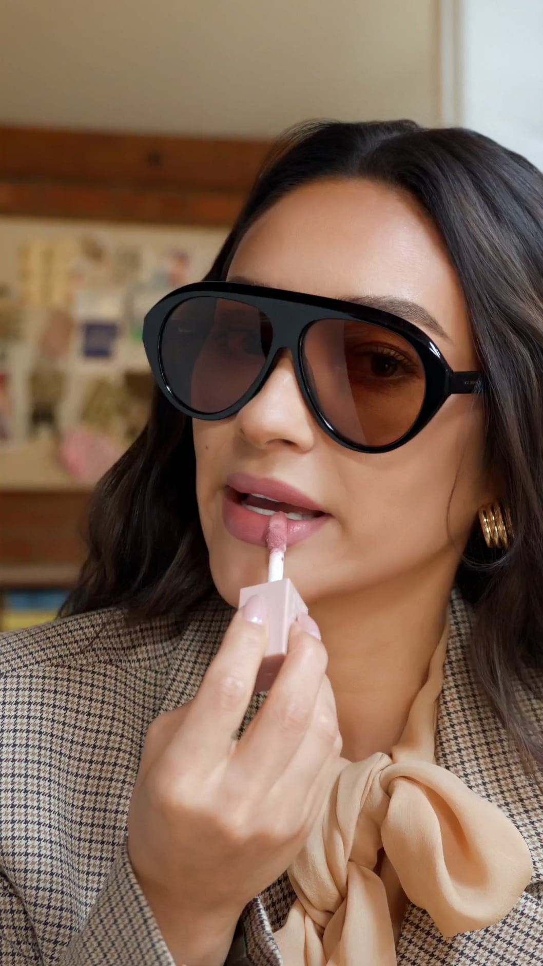 Shay Mitchell in oversized aviator sunglasses applying pink lip gloss, a check blazer and a peach silk bow at her neck.