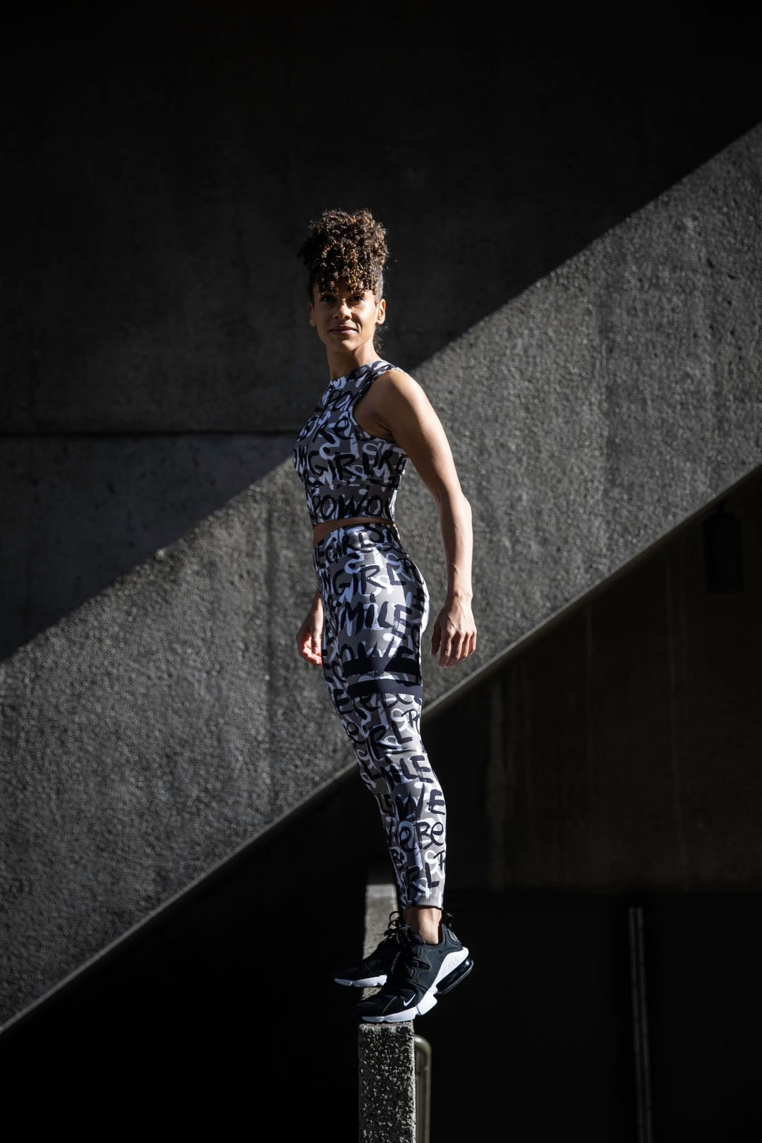 A model in printed activewear standing on a concrete ledge, cut by a hard diagonal of sunlight against deep shadow.