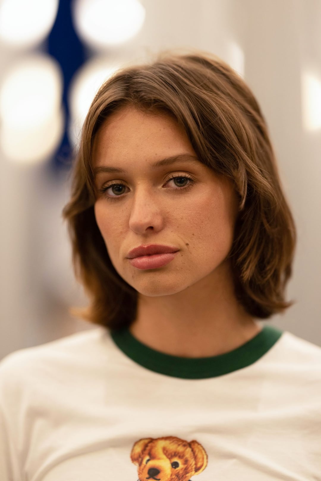 Tatchi close-up in a white ringer tee, soft bokeh of lamp posts behind her, looking into the lens.