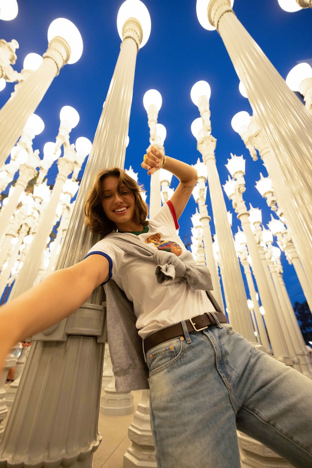 Tatchi seen from below among the lit lamp posts of Urban Light at dusk, one arm flung out, in jeans and a white tee.